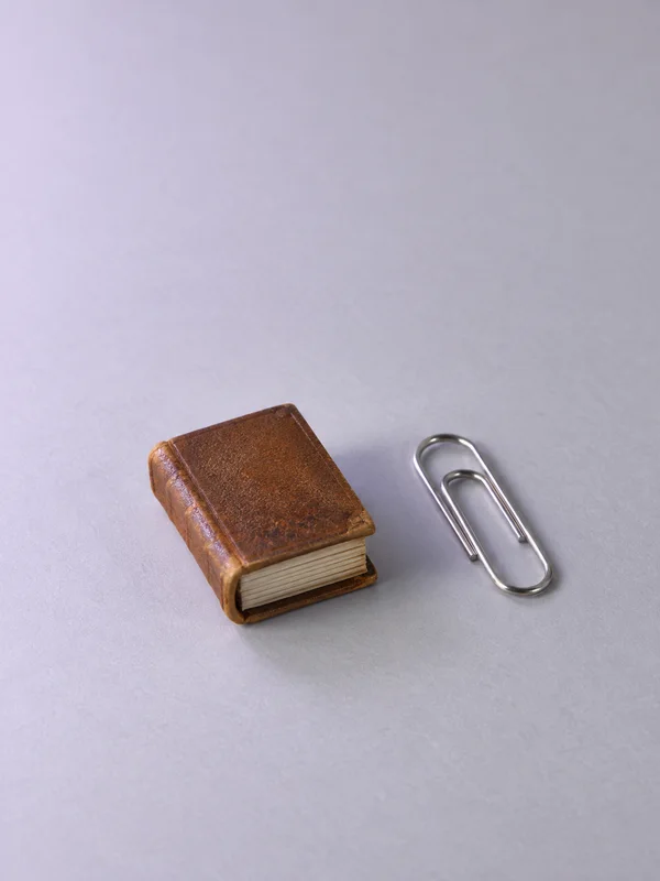 A small closed brown leather book with a cream page block sits beside a silver paper clip on a pale lavender-grey surface, with broad empty space above.