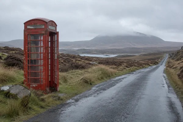 A weathered red telephone box with its door ajar stands beside a wet lane crossing open moorland, with a misty hill and pale grey sky in the distance.