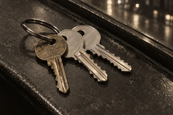 Three metal keys on a split ring lie in a diagonal fan on a dark, textured sill, with warm points of light reflected in the glass behind them.