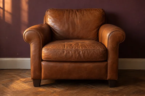 A caramel-brown leather club chair with rounded arms and dark wooden feet sits on oak parquet against a plum wall, lit warmly from the left.