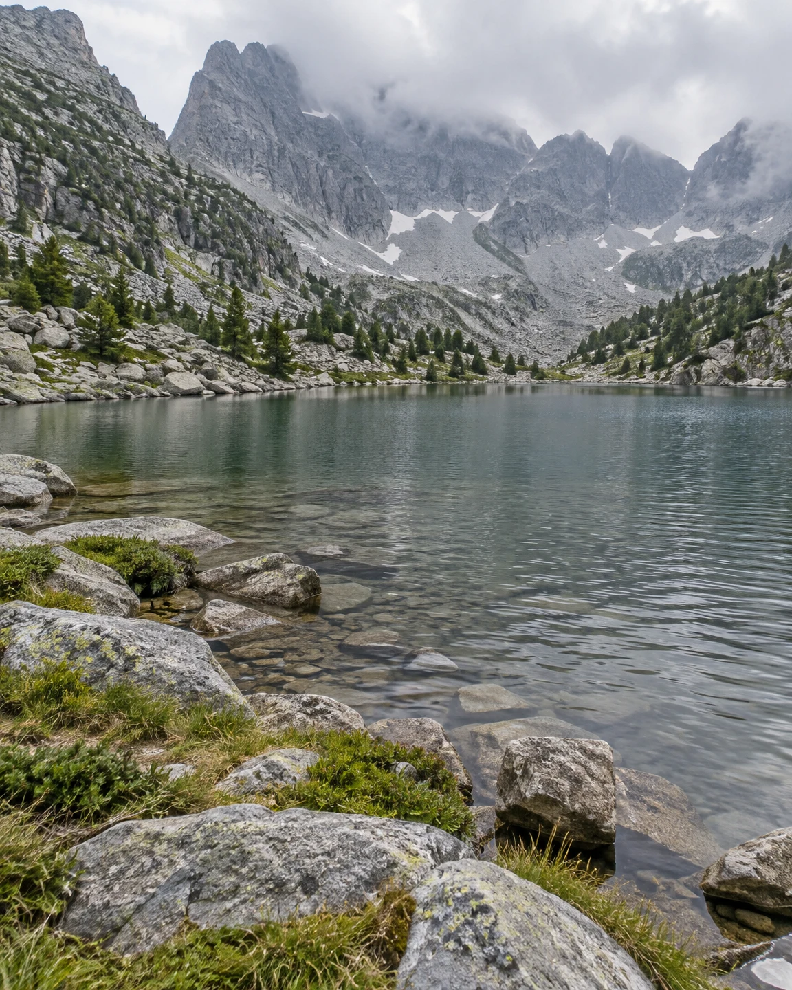 A clear alpine lake fills a rocky mountain cirque, with large granite shoreline stones and grasses in the foreground, rippled water, scattered conifers, and…