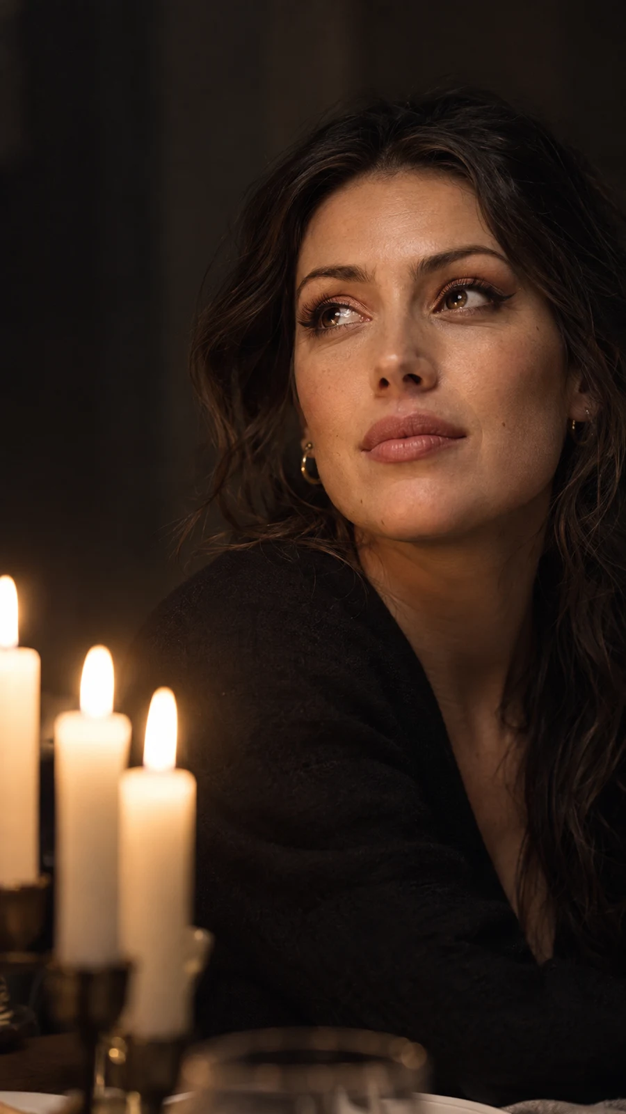 A woman in dark clothing looks slightly upward beside three softly blurred candles in a dark interior, with warm light across her face and copper-toned eye…