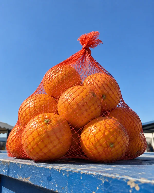 A knotted orange mesh bag filled with oranges rests on a weathered blue-painted counter beneath a clear blue sky, with market roof edges visible in the…