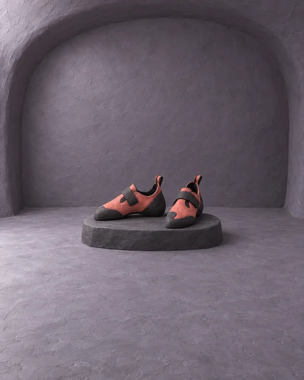 A pair of coral-colored clay climbing shoes with dark soles and fastening straps sits on a low charcoal display platform inside a spacious violet-gray alcove.