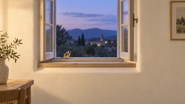 A small goldfinch with a red face and yellow wing stripe perches on the sill of an open timber window in a warmly lit plaster room, with a softly blurred…