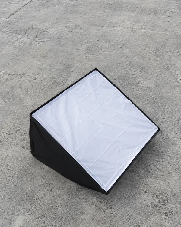 A square black softbox rests on mottled pale-grey concrete, its white, creased diffuser facing upward at a diagonal with one black side panel visible.