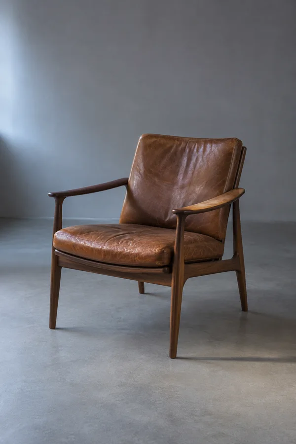 A walnut armchair with a worn brown leather seat and back sits alone on a matte grey studio floor, shown in three-quarter view.