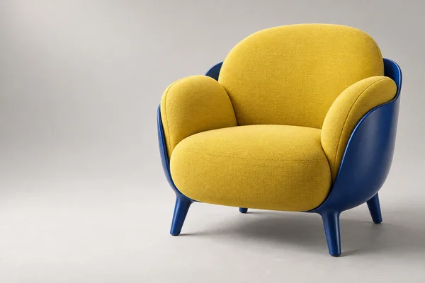 A sculptural armchair with saturated yellow upholstered cushions and a cobalt-blue outer shell and legs sits on a pale warm-grey studio floor, with open space…