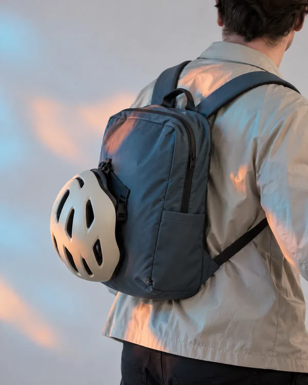 A person seen from behind wears a stone-coloured jacket and slate-blue backpack, with a cream bicycle helmet clipped to the backpack.