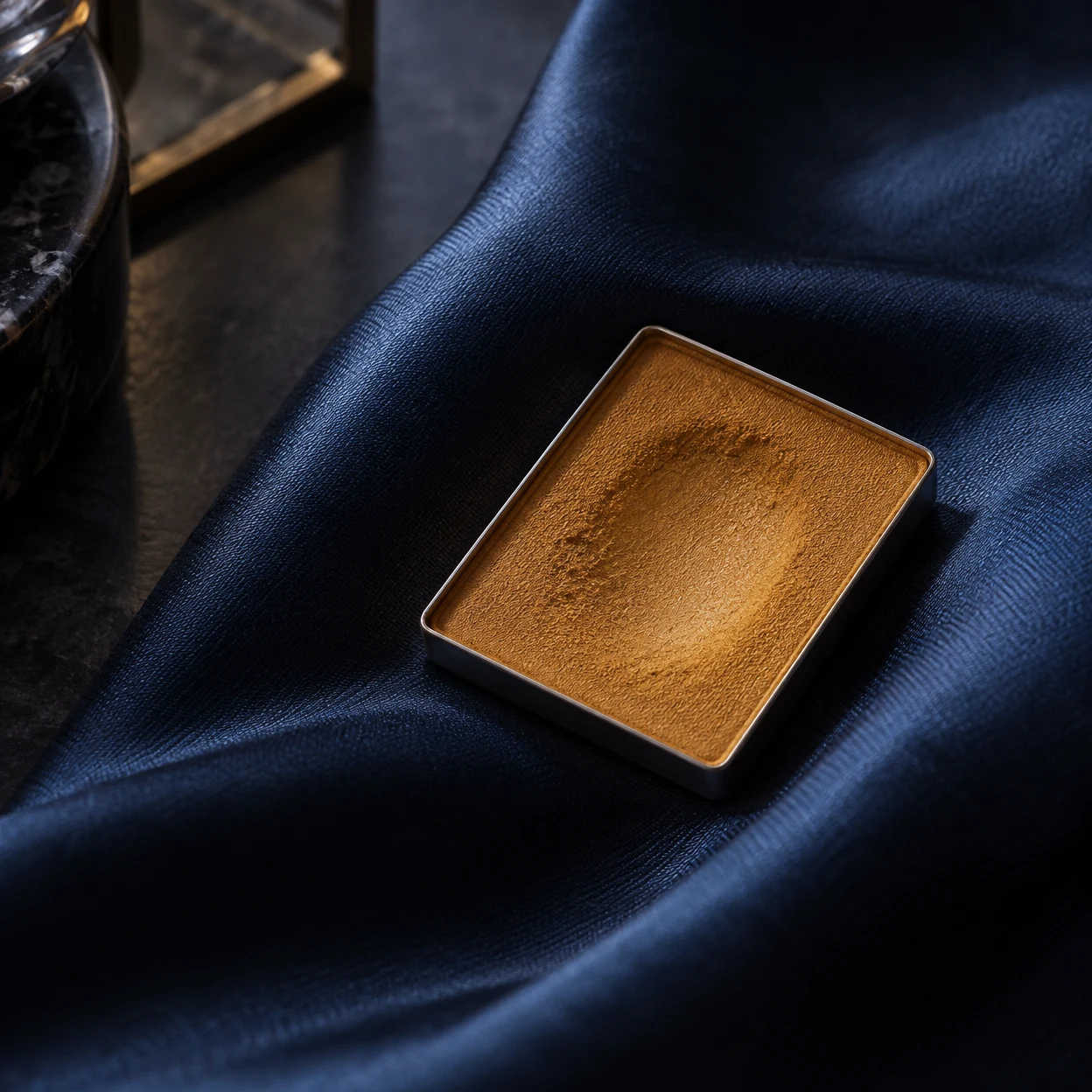 A shallow rectangular metal pan of warm ochre powder rests at an angle on deeply folded indigo satin, with a worn hollow across the powder and cropped dark…