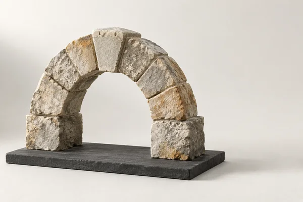 A rough limestone block arch with a prominent central keystone stands on a charcoal slab against an ivory studio background, with open space to the right.