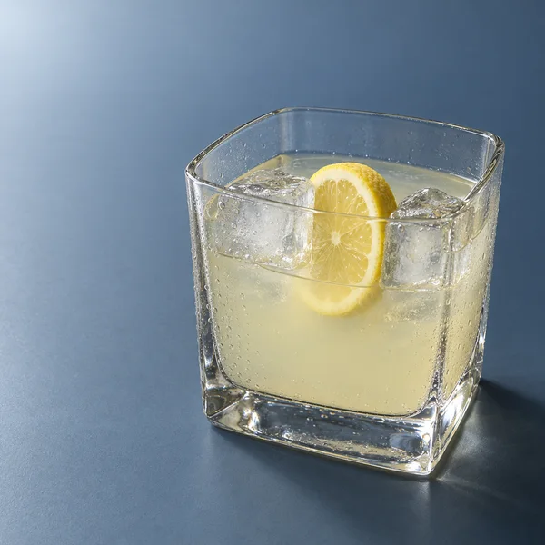 A square heavy glass tumbler of pale yellow lemonade sits on a blue-gray tabletop, with two clear ice cubes flanking an upright lemon wheel and condensation…
