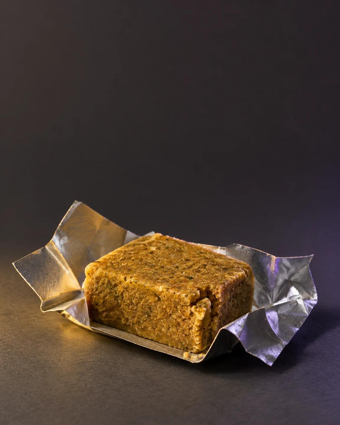 A golden-brown bouillon cube rests in an opened silver foil wrapper on a dark surface, against a charcoal-grey background with warm amber light and violet…