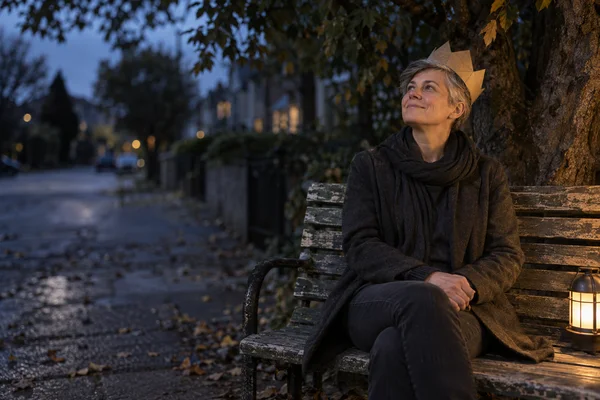 A short-haired adult in charcoal clothing sits on a weathered park bench beneath a tree at dusk, wearing a folded kraft-paper crown and looking up toward the…