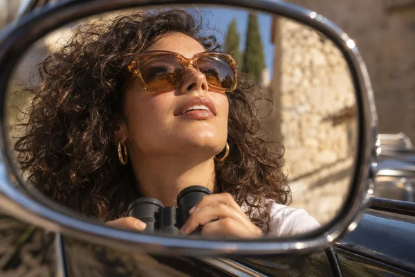 A woman with tousled dark curls, amber sunglasses and gold hoop earrings holds compact black binoculars beneath her chin, looking upward in the reflection of a…