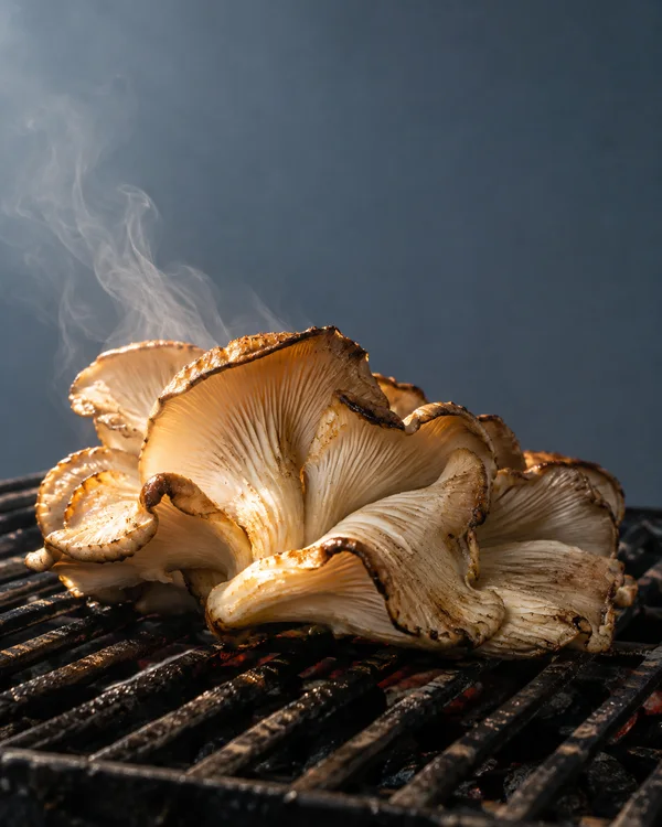 A large cluster of golden-brown oyster mushrooms sits on blackened grill bars against a plain blue-grey background, with pale steam rising above it.