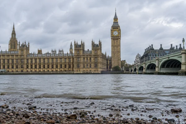 A low view across the Thames from a wet shingle foreshore toward Elizabeth Tower and the Palace of Westminster under an overcast sky, with Westminster Bridge…