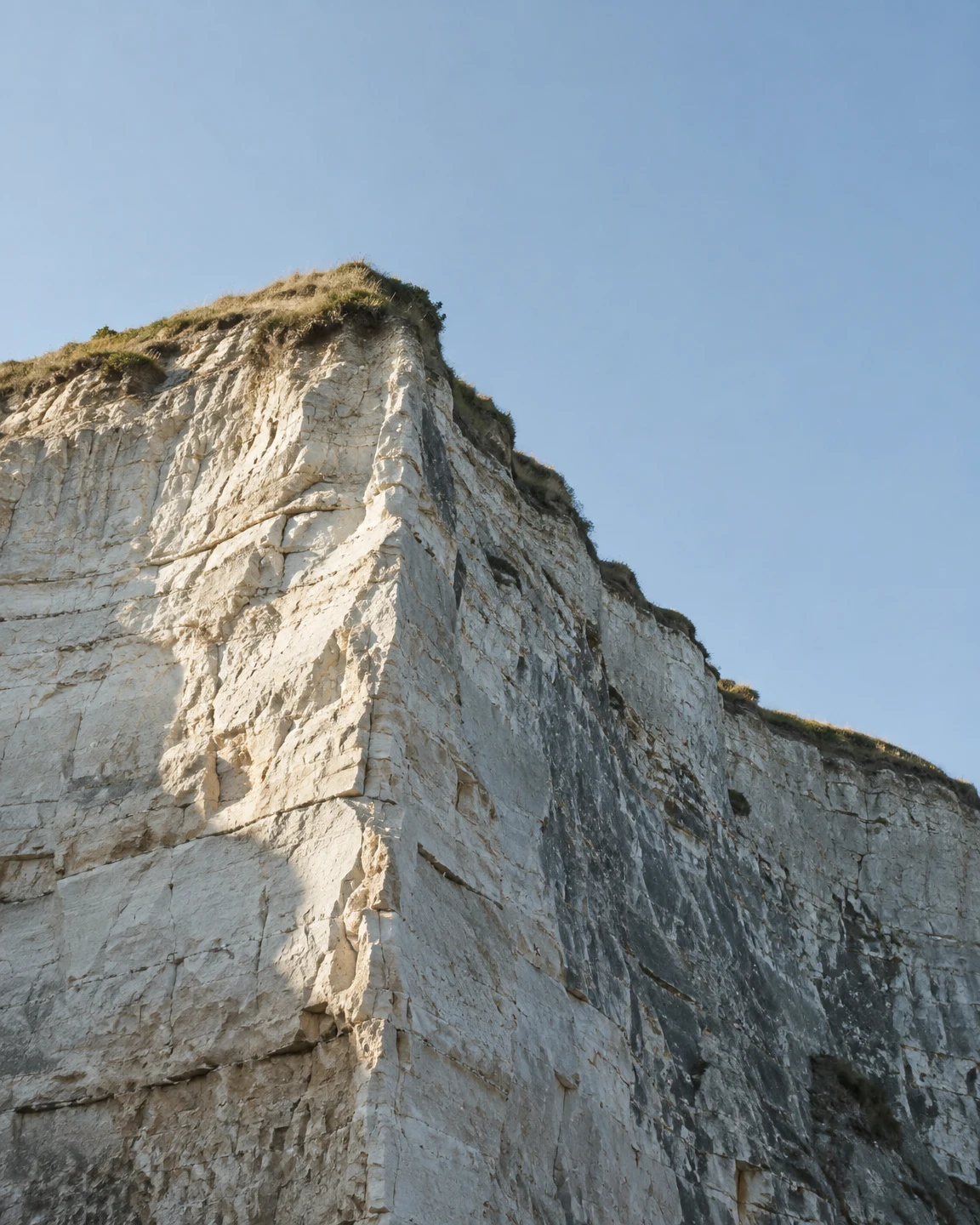 A pale chalk coastal cliff rises beneath a clear blue sky, with warm sunlight on one face and a cool shaded face along its projecting corner; sparse grass…