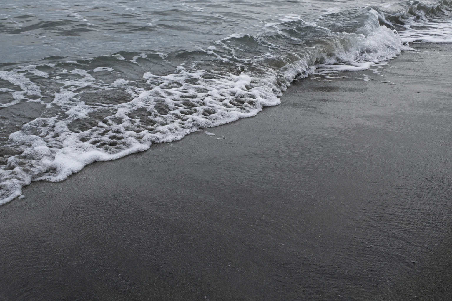 A small grey wave folds onto dark wet sand, with an irregular white foam edge running diagonally across the frame and open sand in the foreground.