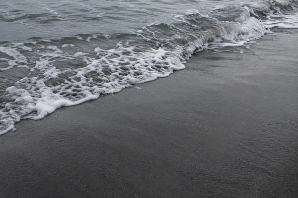 A small grey wave folds onto dark wet sand, with an irregular white foam edge running diagonally across the frame and open sand in the foreground.