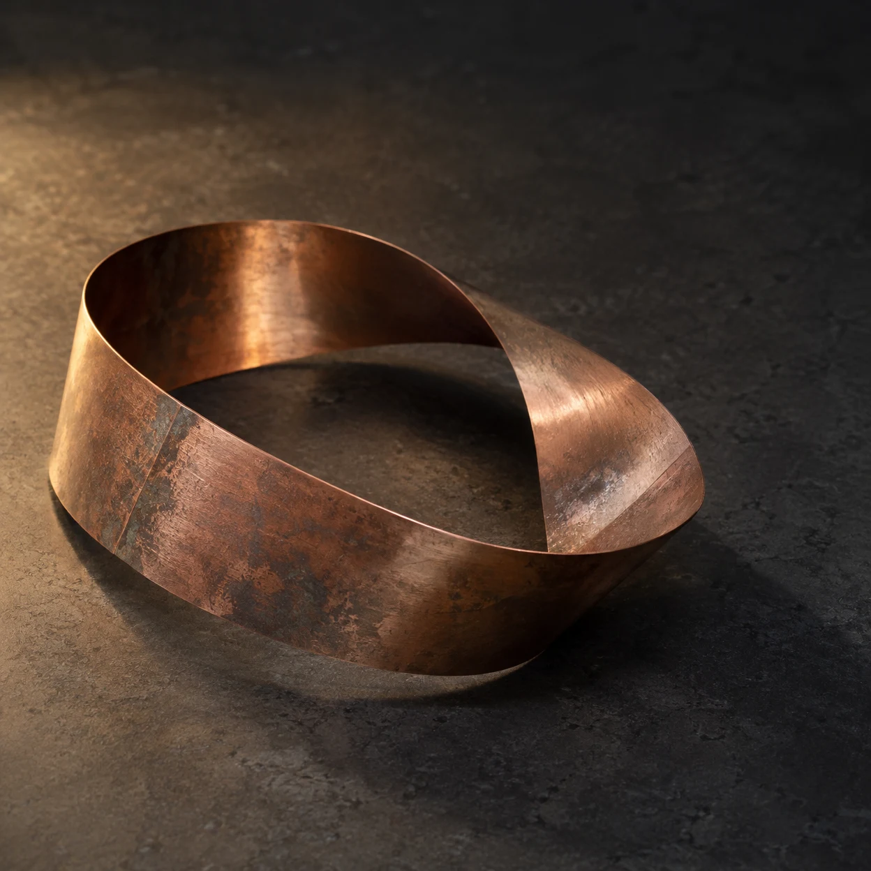 A broad, weathered copper band forms a loop on a dark stone tabletop, lit by a warm pool of light with open space in the upper right.