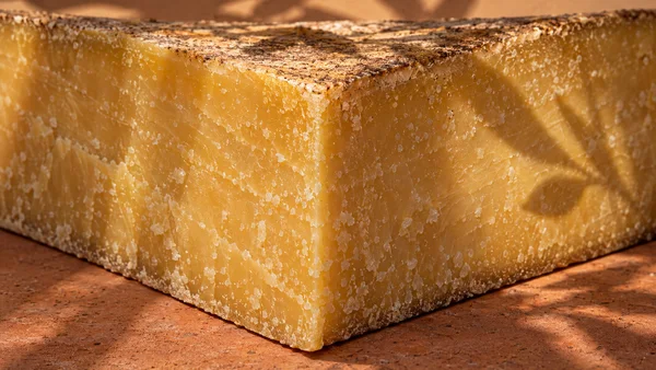 An extreme close-up of a large golden-yellow aged cheese block, its front corner resting on a reddish terracotta tile.