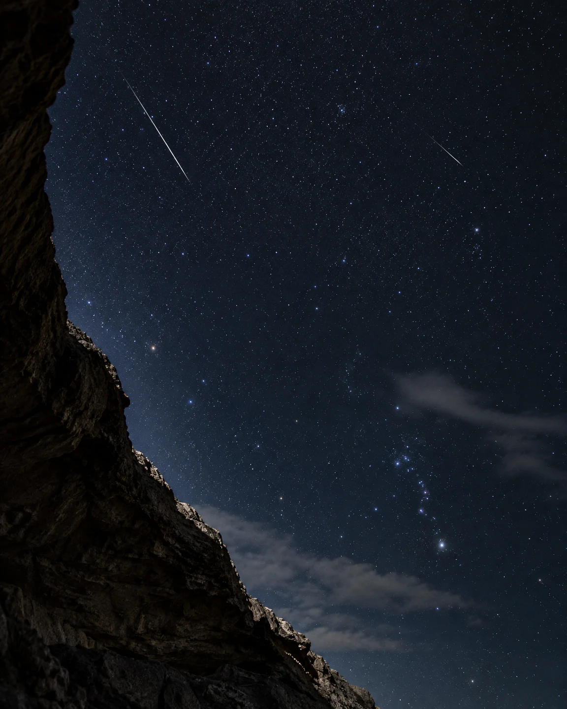 A star-filled deep blue night sky framed by a dark rock shelter on the left, with two thin meteor streaks and wisps of cloud crossing the lower sky.
