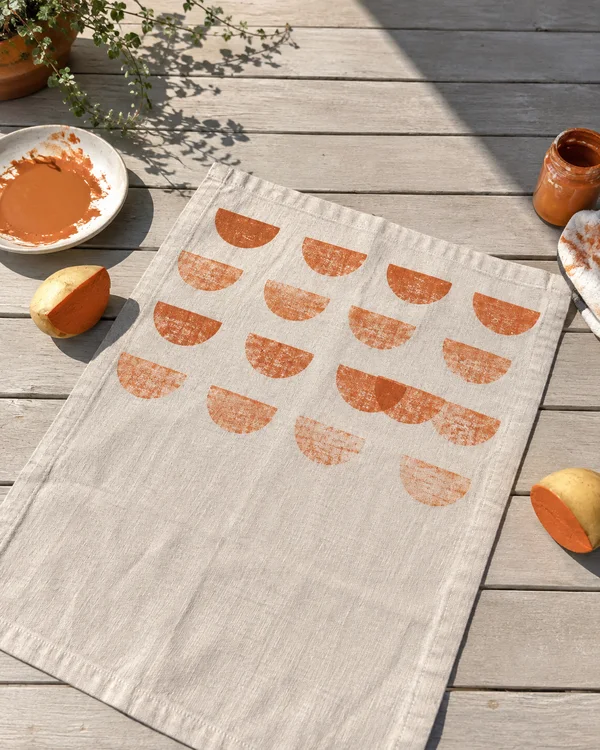 An overhead view of a cream linen towel laid diagonally on a pale wooden table, with loose rows of burnt-orange semicircle prints and a blank lower section.