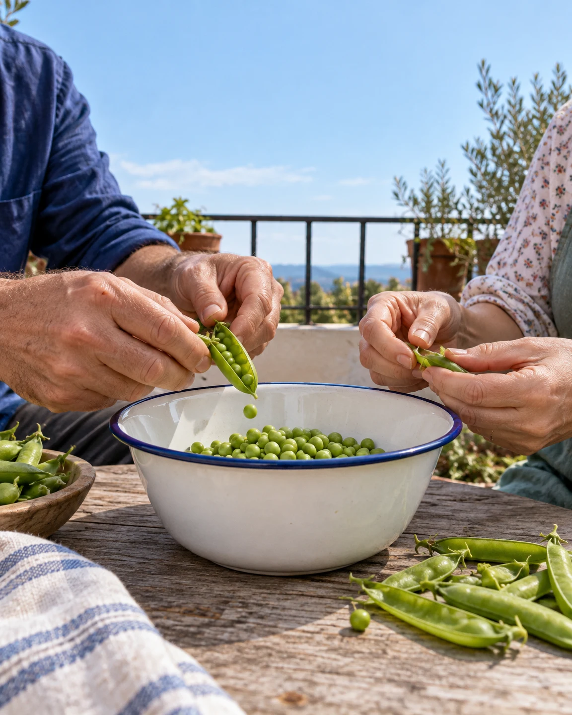 Two cropped people shell fresh peas over a large white enamel bowl with a dark blue rim on a wooden patio table.