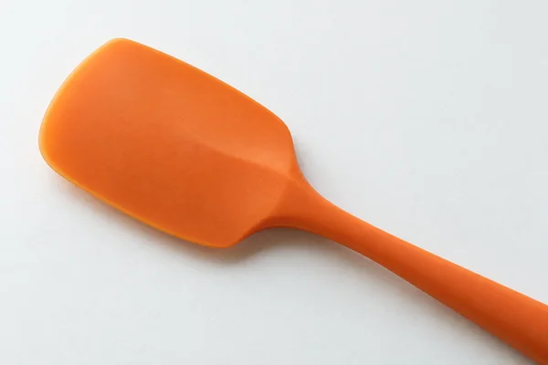 A tangerine-orange silicone spatula lies face-up on an off-white surface, with its broad rounded head on the left and handle extending diagonally beyond the…