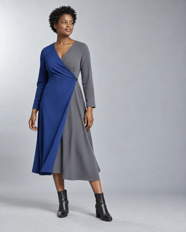 A woman with short natural curls wears a cobalt-blue and slate-gray wrap dress with a button fastening at the hip and black ankle boots, posed against a pale…