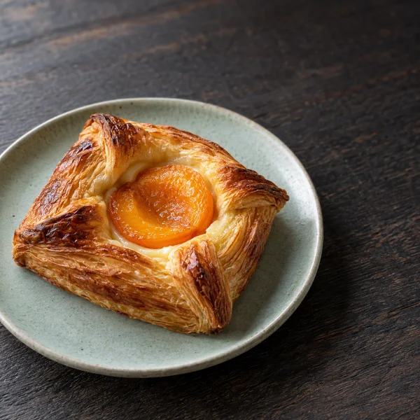 A golden apricot Danish with pale cream sits on a pale celadon plate on a dark wooden table, with open tabletop in the upper right.