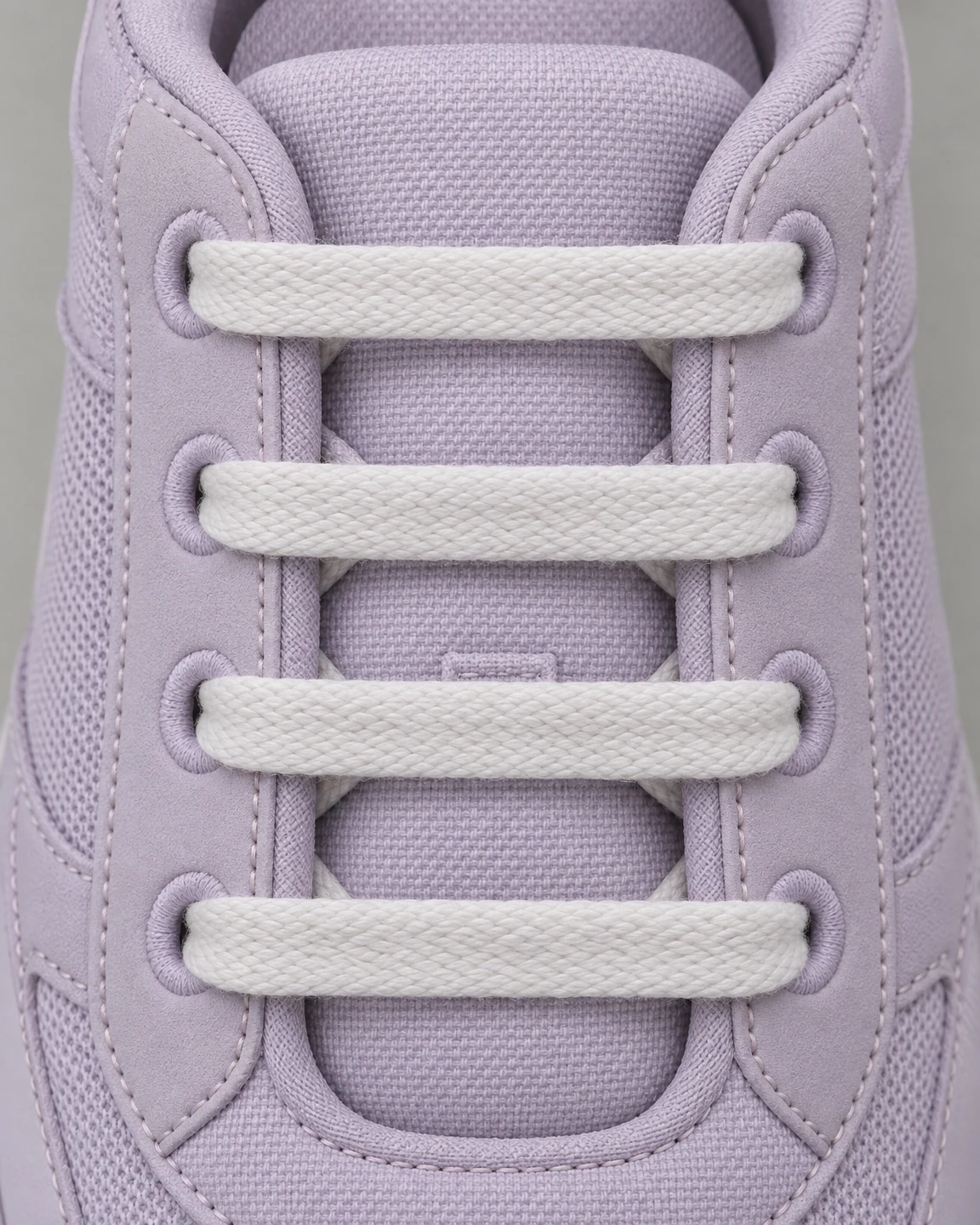 Overhead close-up of four broad white laces crossing the padded tongue of a pale lilac walking shoe, with fabric eyelets and stitched panels visible against a…