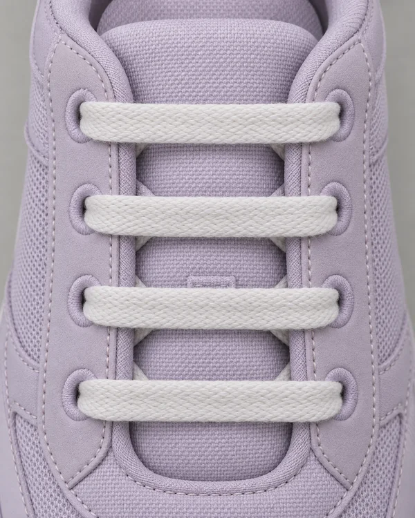 Overhead close-up of four broad white laces crossing the padded tongue of a pale lilac walking shoe, with fabric eyelets and stitched panels visible against a…