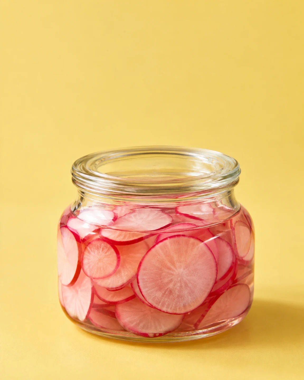 A squat clear glass jar filled with overlapping radish rounds in pink brine sits on a butter-yellow surface against a matching background, with generous empty…