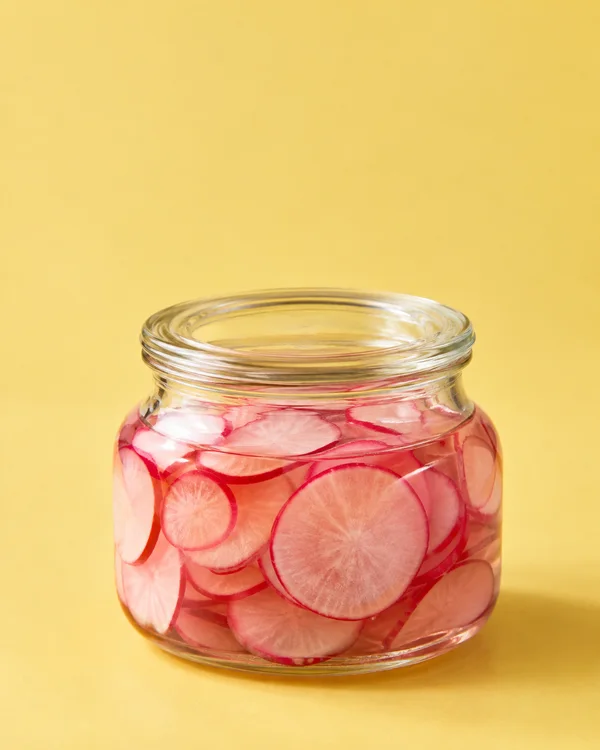 A squat clear glass jar filled with overlapping radish rounds in pink brine sits on a butter-yellow surface against a matching background, with generous empty…