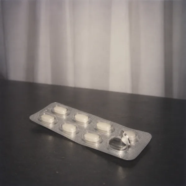 A silver blister pack of ivory tablets lies diagonally on a charcoal tabletop, with one pocket opened and its foil peeled back; soft gray-white curtains fill…