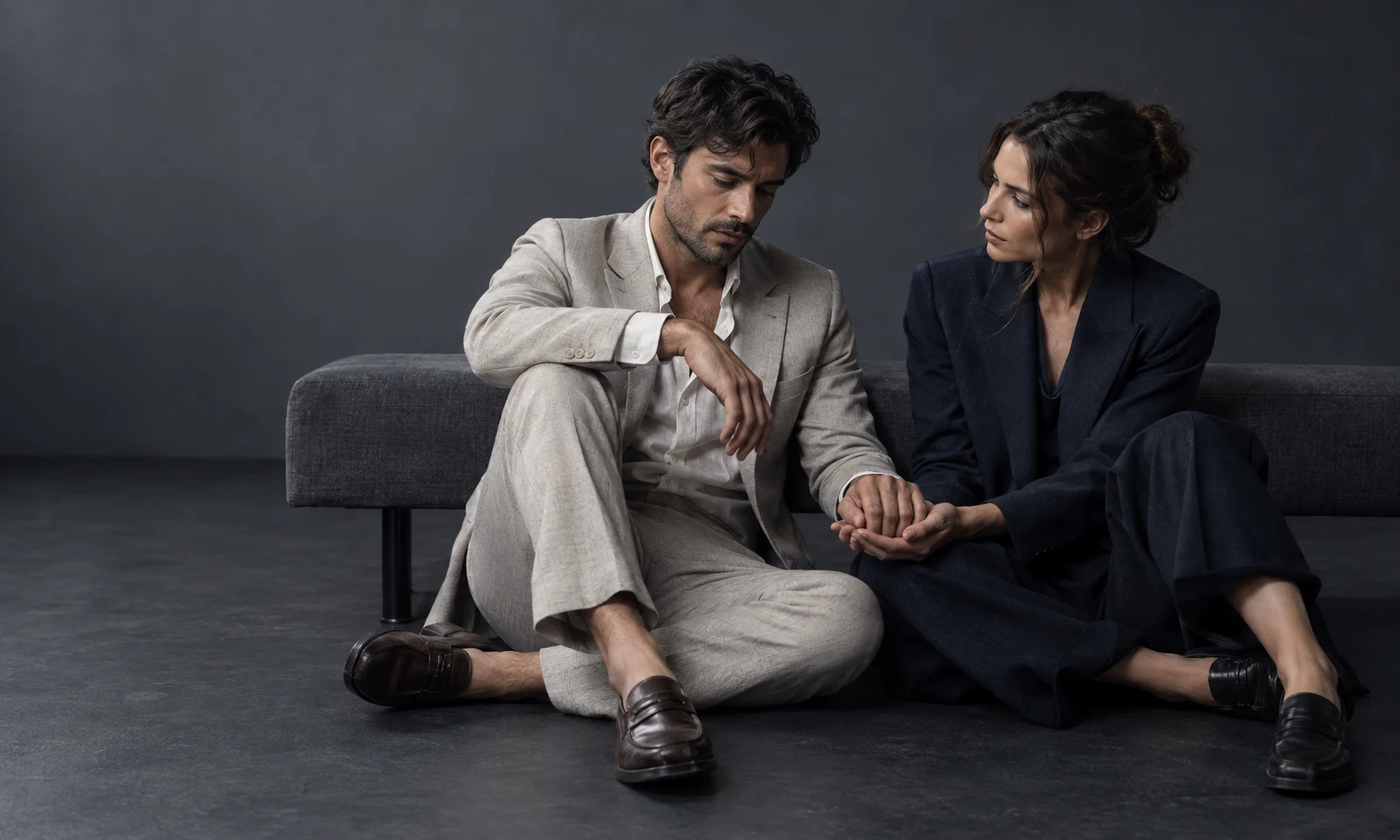 A man in a light stone suit and a woman in dark navy tailoring sit on a charcoal floor in front of a low upholstered bench.