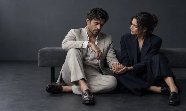A man in a light stone suit and a woman in dark navy tailoring sit on a charcoal floor in front of a low upholstered bench.