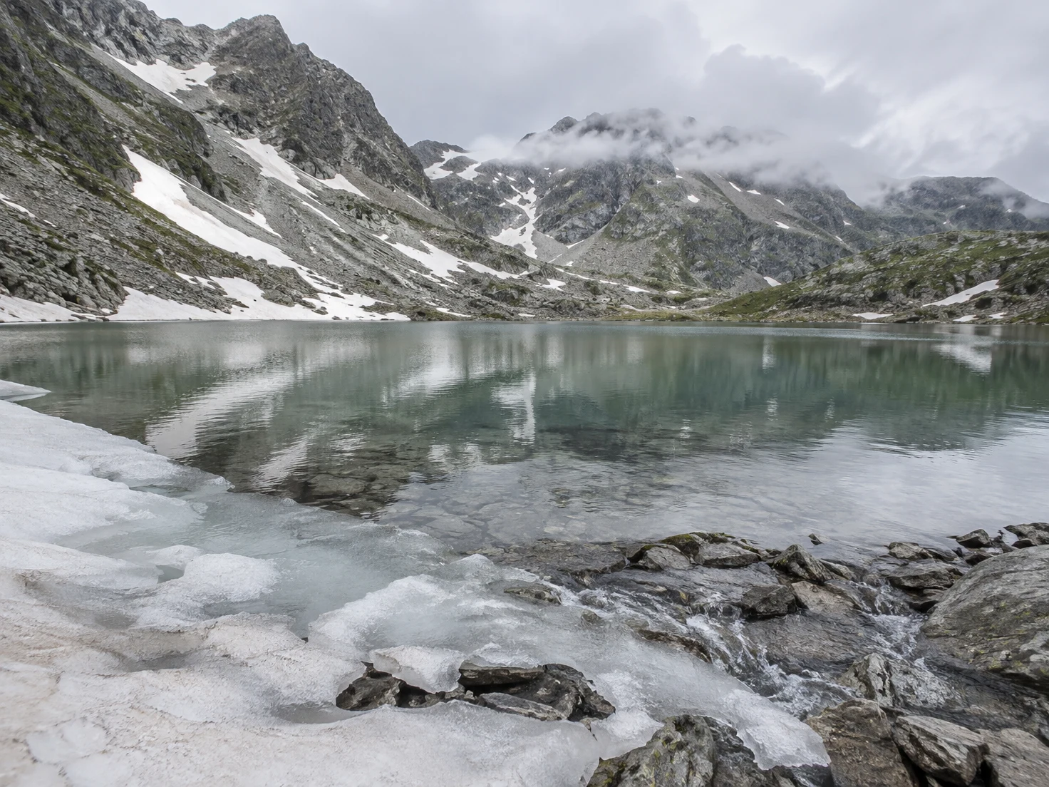 A calm grey-green alpine lake sits beneath rocky, snow-streaked peaks softened by low cloud.