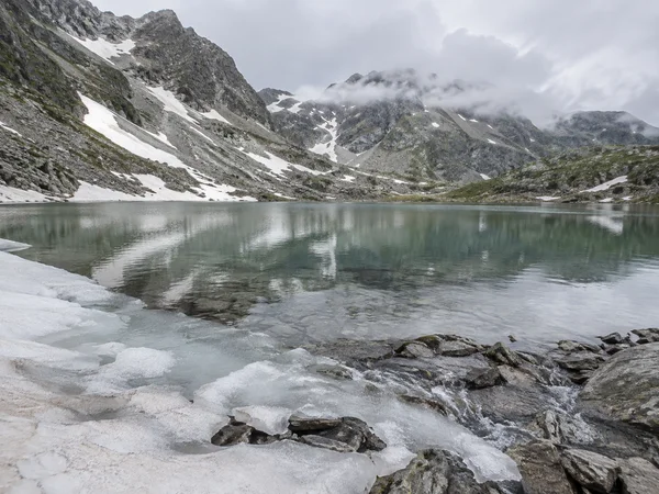 A calm grey-green alpine lake sits beneath rocky, snow-streaked peaks softened by low cloud.