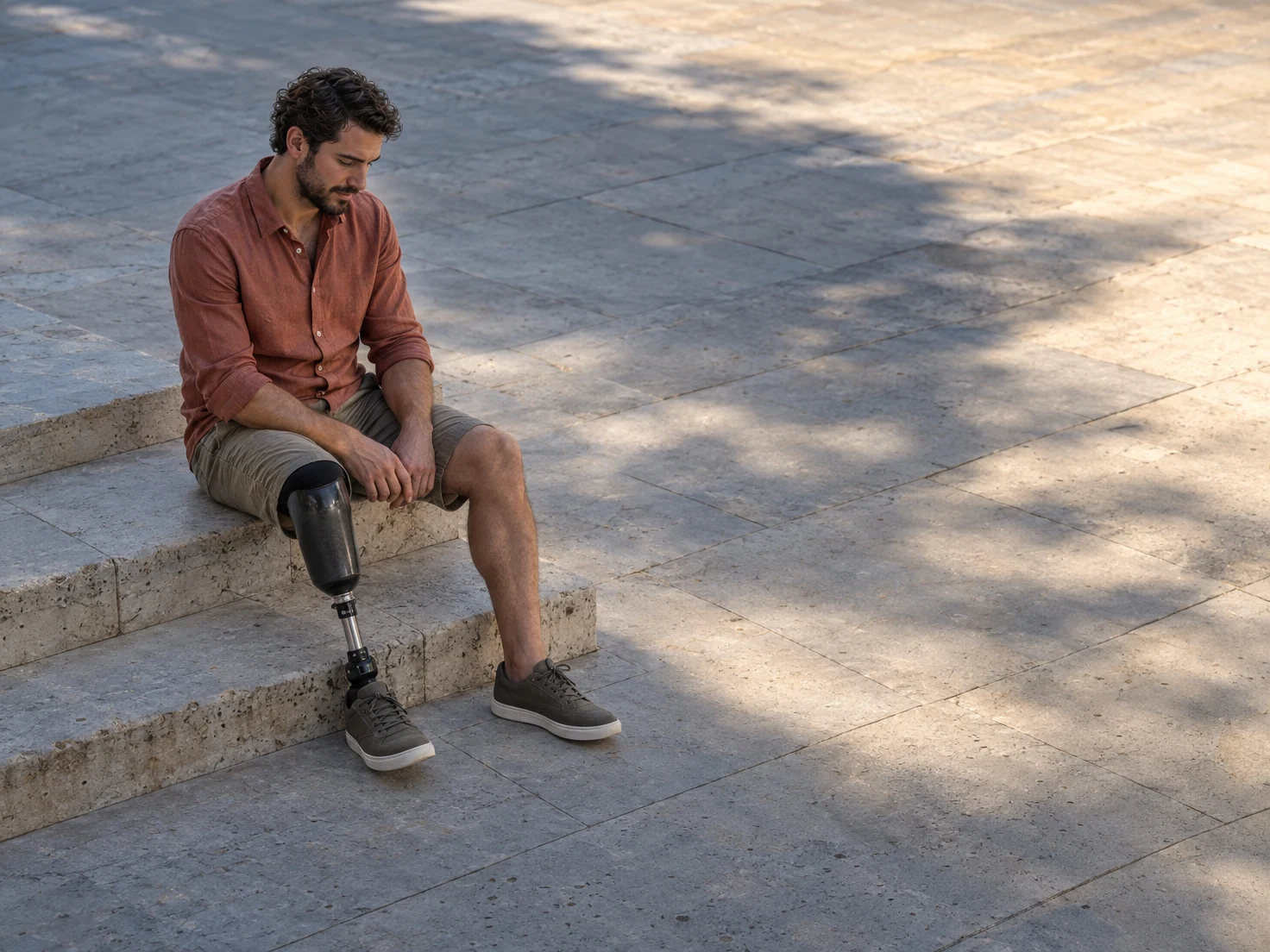 A bearded adult sits with his gaze lowered on limestone steps in an open stone plaza, wearing a terracotta shirt and shorts; a below-knee prosthesis is…