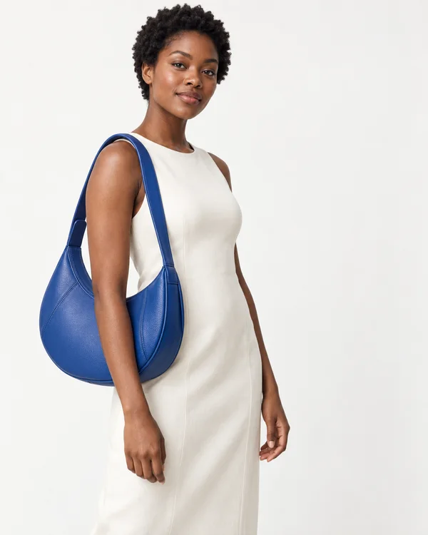 A woman with short natural curls wears an ivory sleeveless dress and carries a large cobalt-blue crescent-shaped leather bag against a warm-white studio…