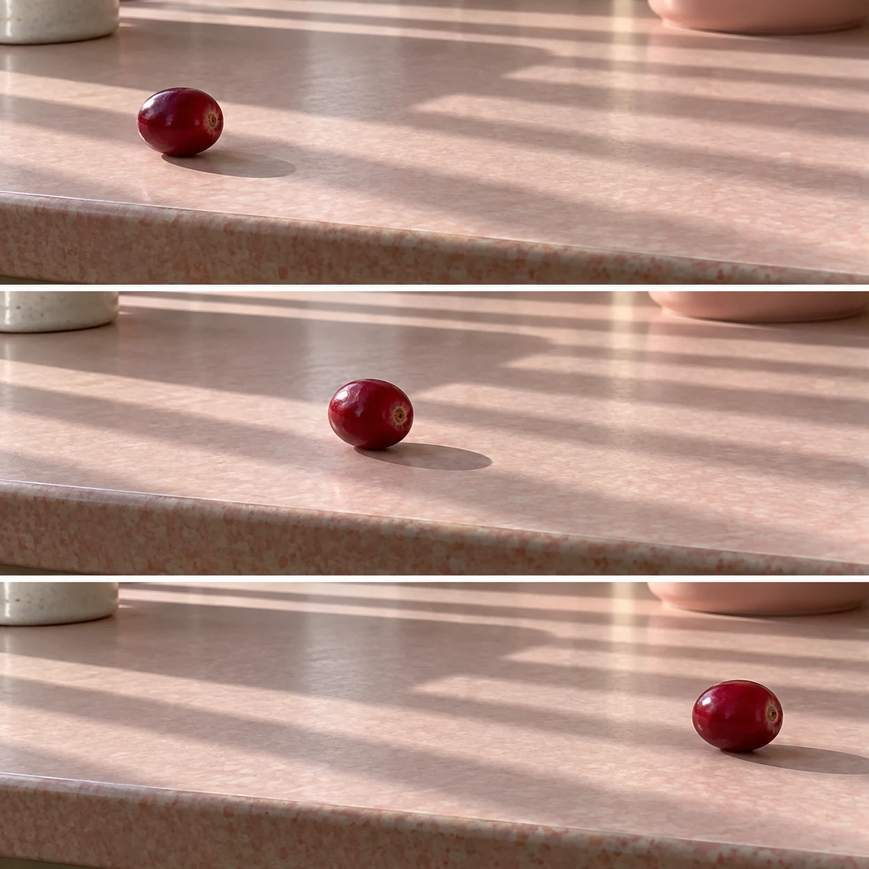 Three horizontal photographs show a crimson cranberry moving from left to right across a pale pink laminate tabletop, with parallel bands of sunlight and…