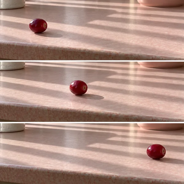 Three horizontal photographs show a crimson cranberry moving from left to right across a pale pink laminate tabletop, with parallel bands of sunlight and…
