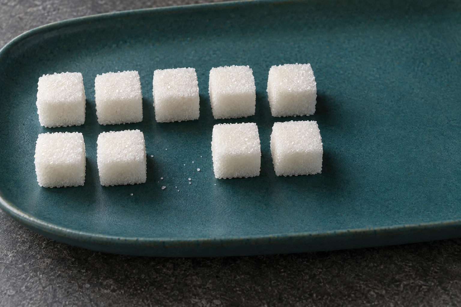 White sugar cubes sit in two rows on a deep-teal ceramic platter, with a gap in the front row and a few loose grains nearby.