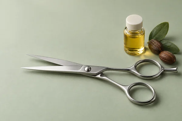 A pair of polished satin-steel haircutting shears lies slightly open on a pale sage surface, with the blades pointing left.