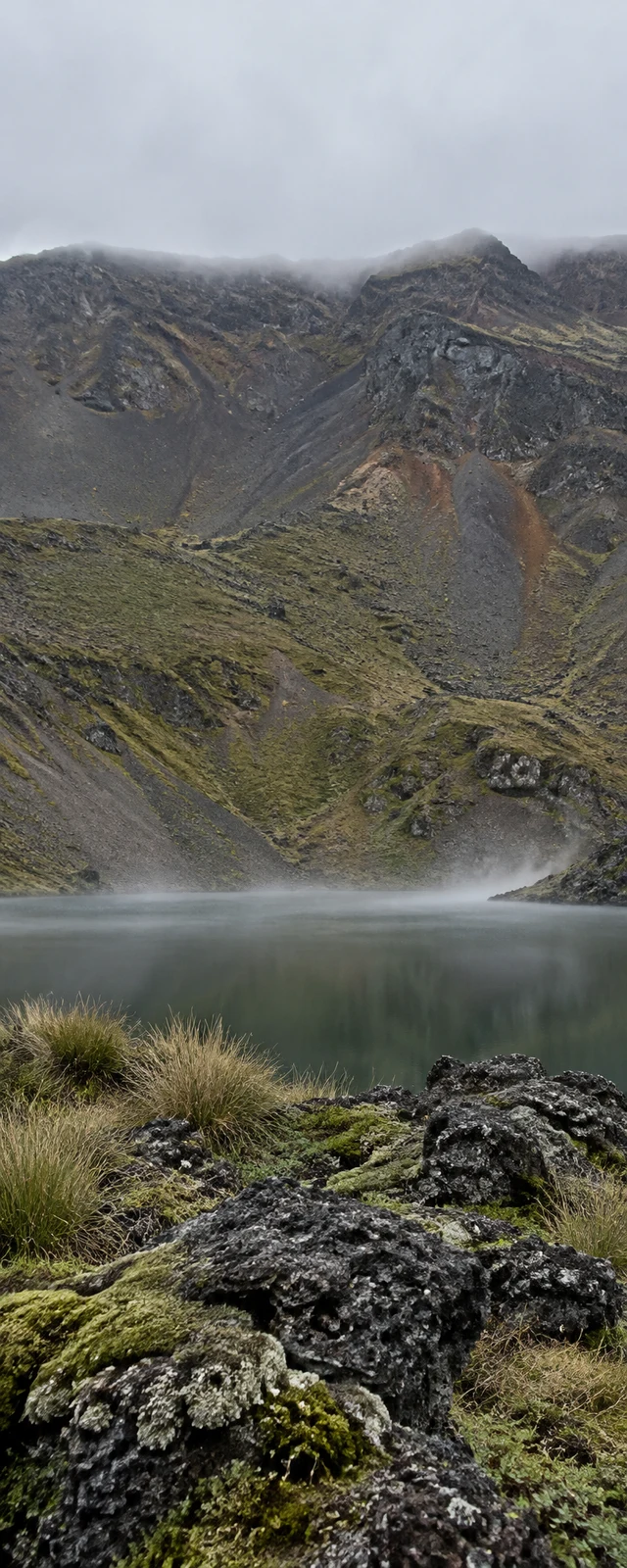 A slate-green volcanic lake lies below a steep, grey and rust-toned rocky ridge under low cloud, with mist hovering over the water and mossy basalt rocks and…