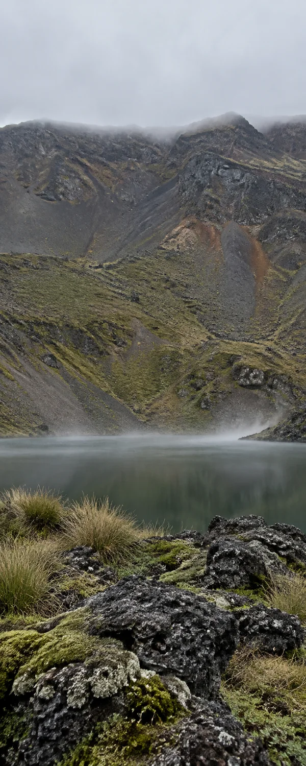 A slate-green volcanic lake lies below a steep, grey and rust-toned rocky ridge under low cloud, with mist hovering over the water and mossy basalt rocks and…