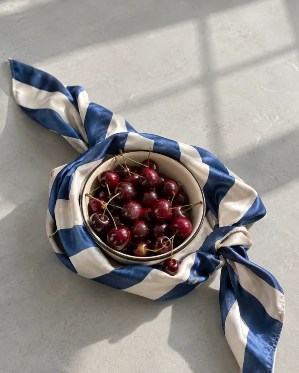 A cream-colored bowl filled with dark red cherries sits in a blue-and-ivory striped silk scarf on a pale grey floor, viewed from above.